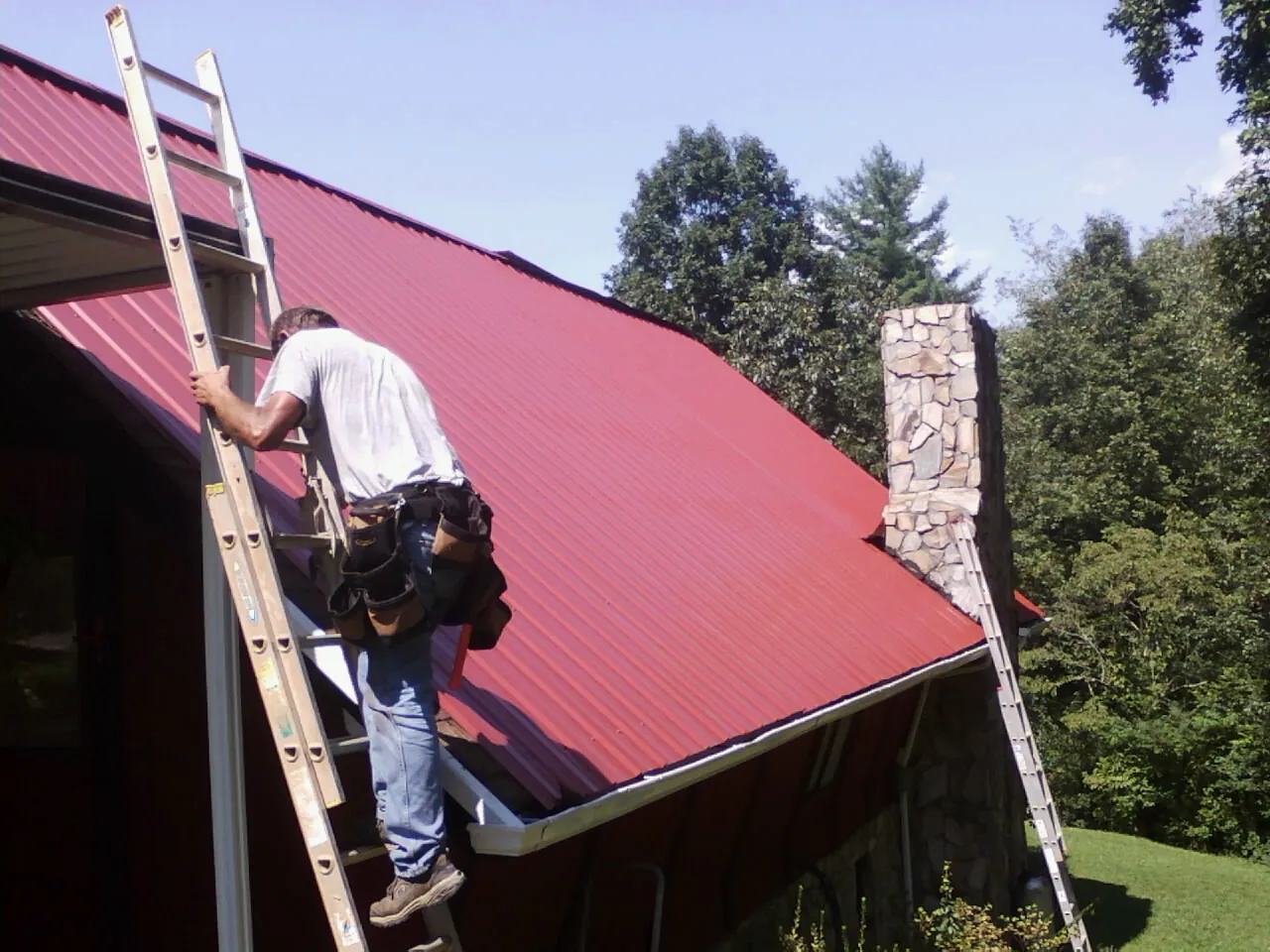 Roofer on a ladder beside a red metal roof and stone chimney — Soffit & Fascia Repair by Roofing Brothers in Willington